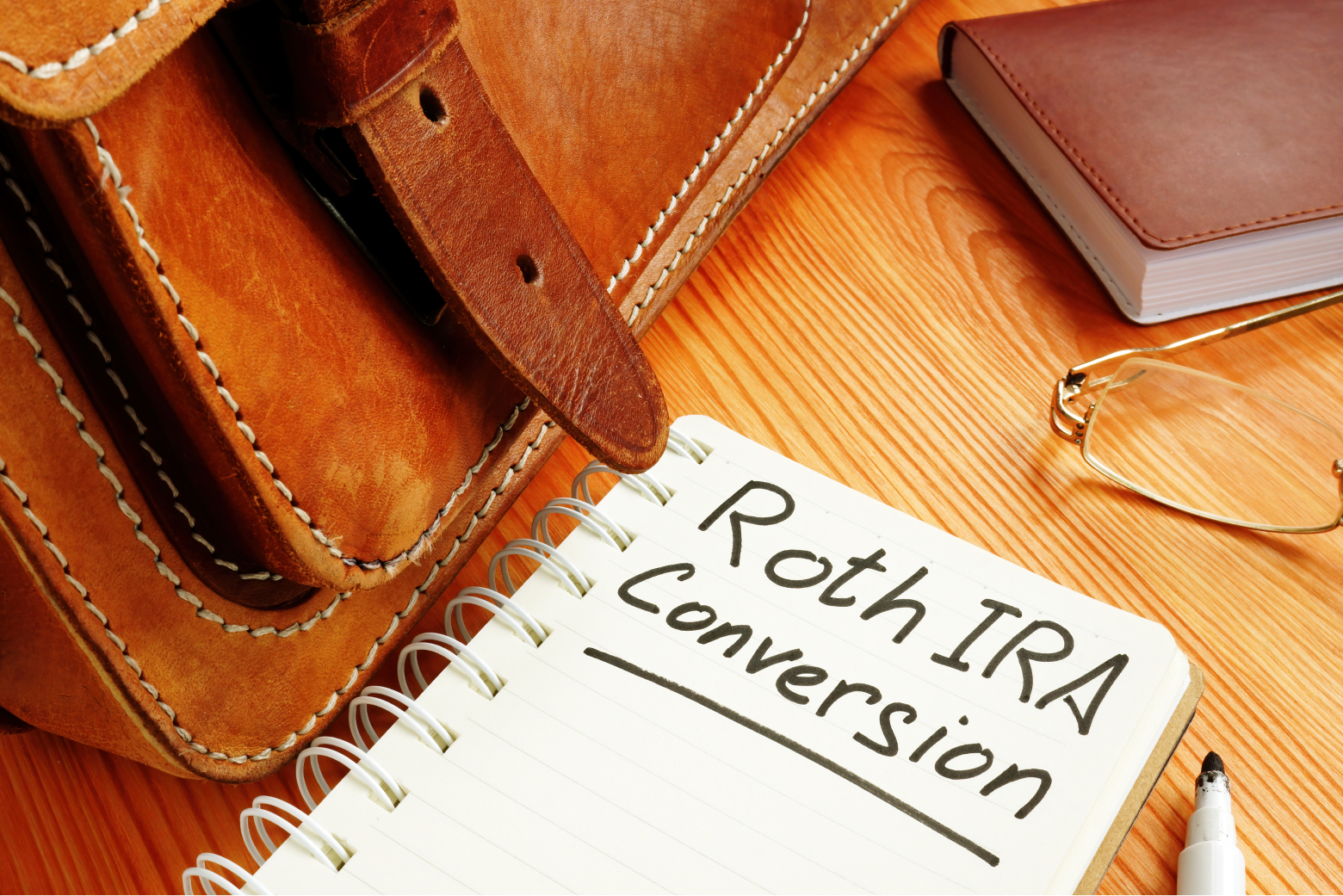 A notebook with Roth IRA Conversion written on it sits on a table in front of a leather messenger bag