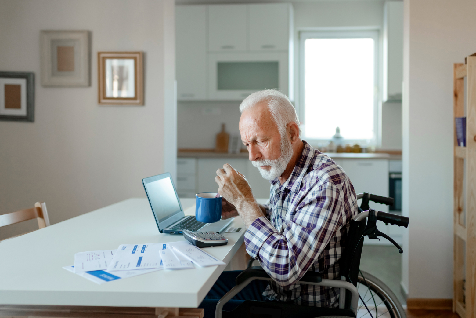 An older white man using a wheelchair sits at a table with a laptop, calculator, and papers.