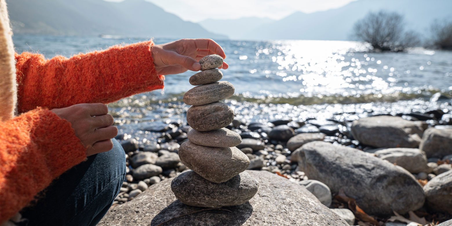 A person places rocks in a tall pile in front of a lake